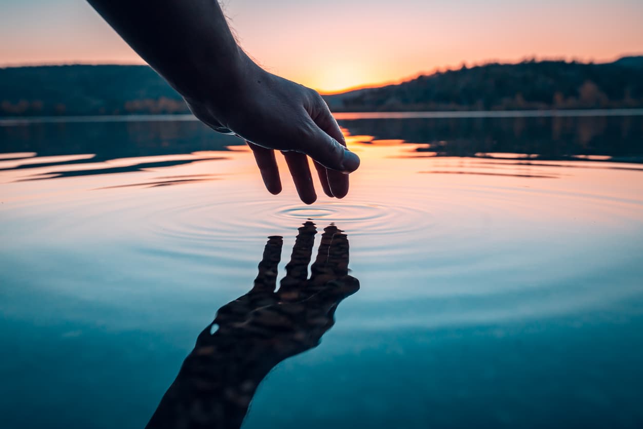 A hand reaching toward its reflection on calm water at sunset